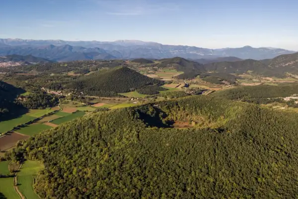 Paisaje de la ruta volcanes Garrotxa, con conos volcánicos cubiertos de bosque y campos verdes, bajo un cielo azul.